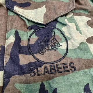 Embroidered "Seabees" insignia of fighting bee logo embroidered in black thread on the left chest pocket of Military BDU Green Woodland Camouflage. Made in U.S.A.